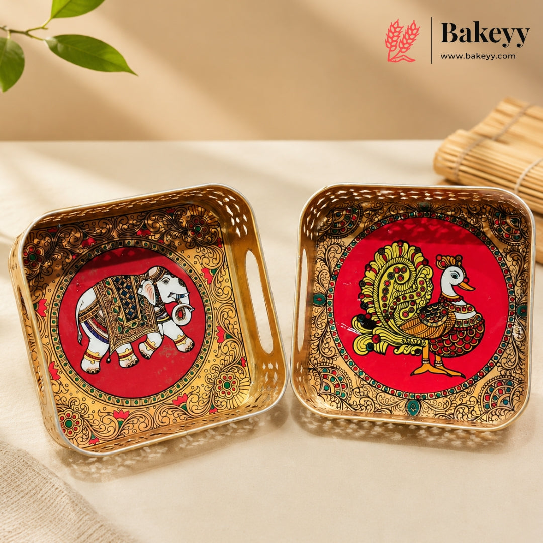 Two decorative square trays with handles, shown on a light beige surface. One tray features a red circular background with a painted elephant, and the other features a painted peacock, both surrounded by ornate gold and multicolor patterns.
