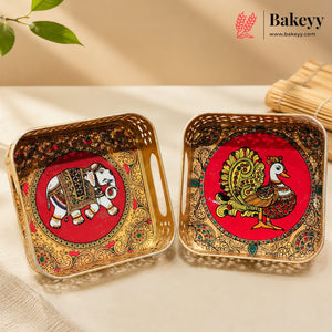 Two decorative square trays with handles, shown on a light beige surface. One tray features a red circular background with a painted elephant, and the other features a painted peacock, both surrounded by ornate gold and multicolor patterns.