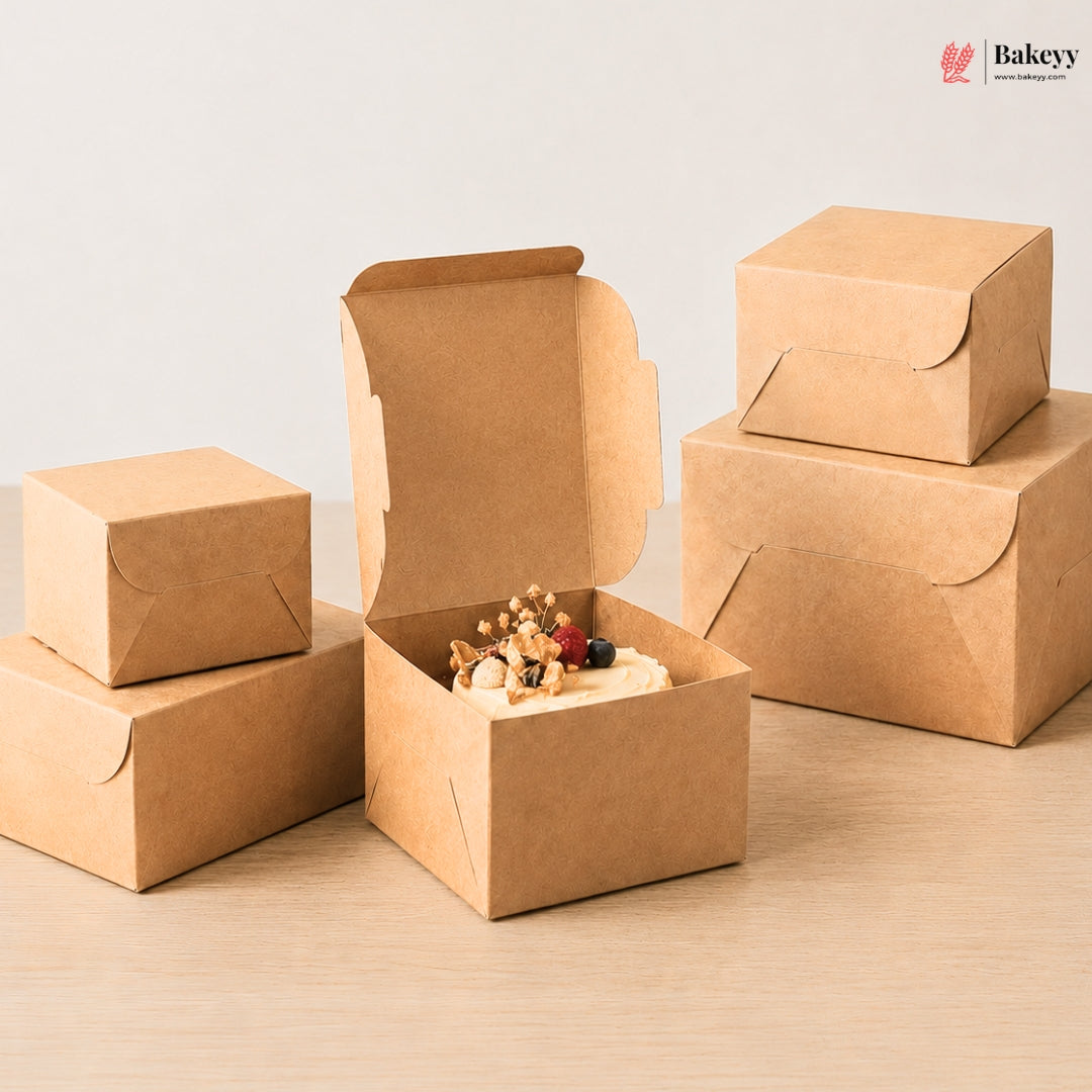 A set of small brown kraft paper bakery boxes in different sizes, with one open box in the center containing a decorated dessert, on a light wood surface against a plain light background. A Bakeyy logo appears in the top right corner.