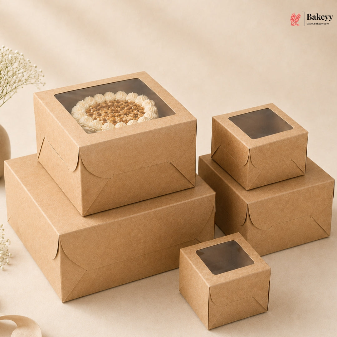 A set of kraft brown bakery boxes with clear window lids, shown in multiple sizes. One larger box is stacked on top of another, holding a round cake with white frosting and nuts.