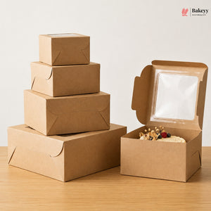 A set of brown kraft paper bakery boxes on a light wood table against a white wall, with one stack of closed boxes on the left and one open box on the right containing a slice of cake with a clear window.