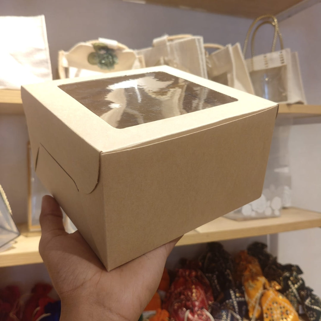A hand holding a square kraft paper gift box with a clear plastic window on top, set against a background of shelves with bags and fabric items.