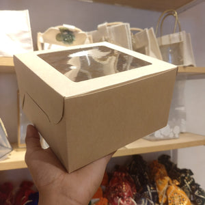 A hand holding a square kraft paper gift box with a clear plastic window on top, set against a background of shelves with bags and fabric items.