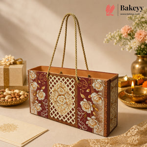An ornate rectangular gift box with rope handles, decorated with cream floral and lattice patterns on a deep burgundy background, styled on a beige surface with sweets, flowers, and a lit brass lamp in the background. The Bakeyy logo is visible in the top right corner.