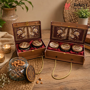 Two ornate brown wooden gift boxes with floral inlay and red velvet lining, each holding three round jars, displayed on a wooden table with candles, a glass jar of almonds, and decorative props. The Bakey brand logo appears in the top right corner.