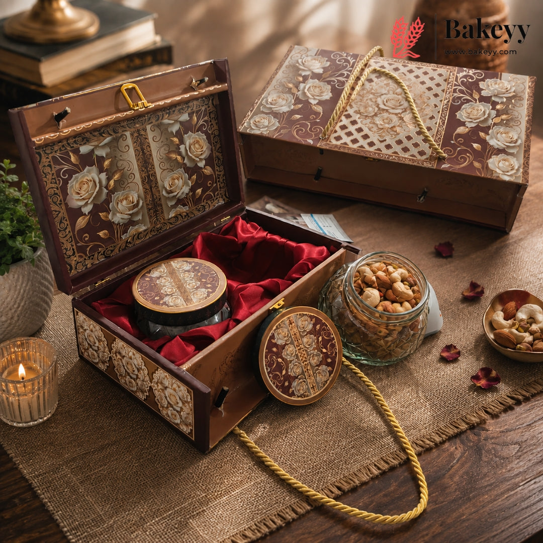 An ornate decorative wooden gift box with floral embossed panels and a red velvet interior, shown open with a matching round tin inside. A second closed box, a glass jar of nuts, and small bowls of nuts are arranged on a rustic table with a candle, books, and scattered rose petals.