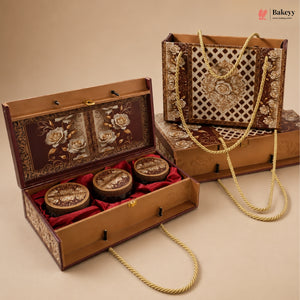 An ornate rectangular gift box set with a hinged wooden box shown open, containing three round decorative jars on red velvet lining, alongside a matching tote bag with rope handles and a closed box. The packaging features gold floral and geometric patterns on a dark brown background, photographed on a beige studio backdrop.