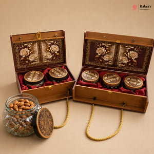 Two ornate wooden gift boxes with floral embroidery and red velvet lining, each holding three round jars, displayed on a beige background with a glass jar of almonds in the foreground. The Bakeyy brand logo is visible in the top right corner.