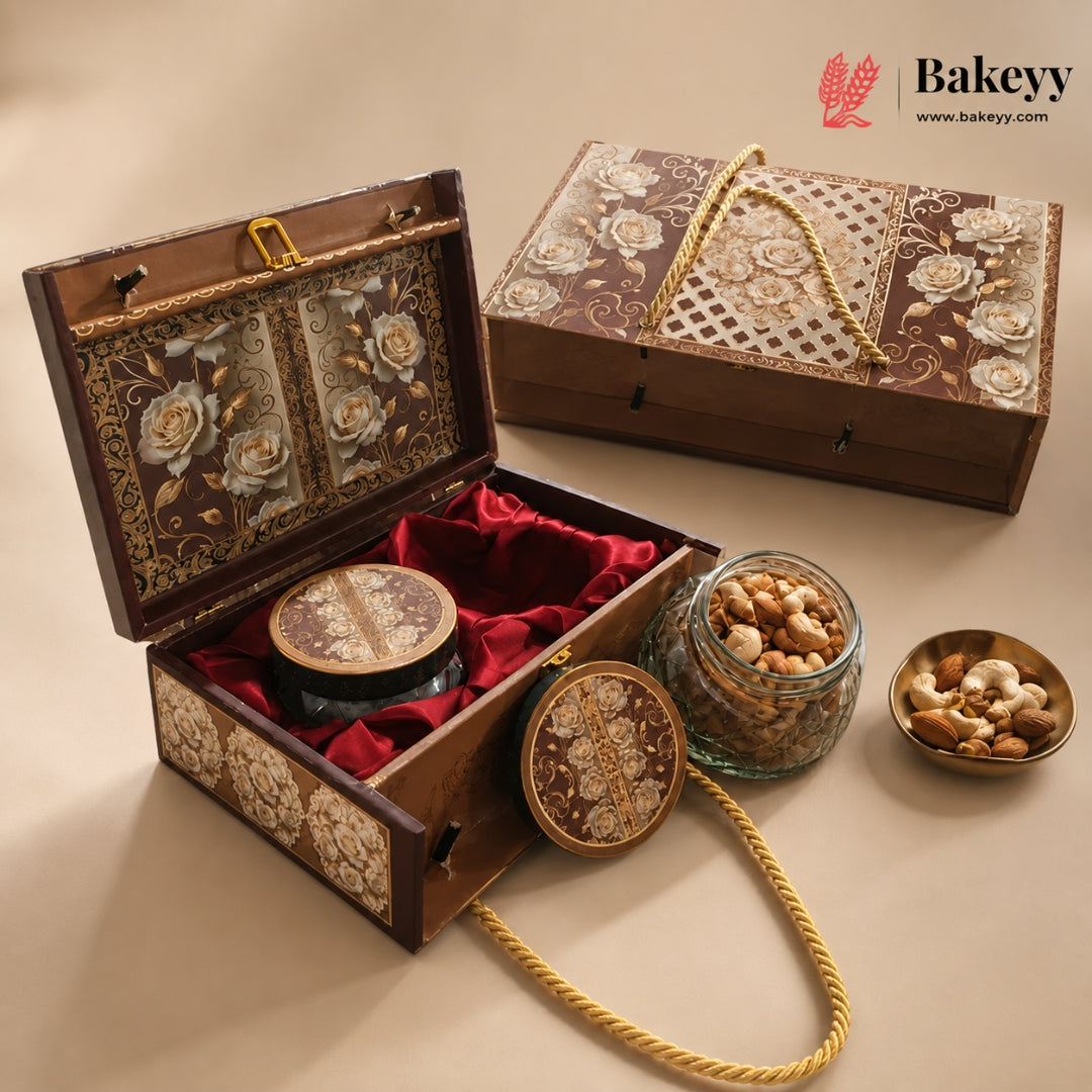 An ornate wooden gift box set with floral inlay, red velvet lining, and gold rope handles, shown open with a round tin and jars of nuts beside it on a beige studio background. The Bakeyy logo appears in the top right.