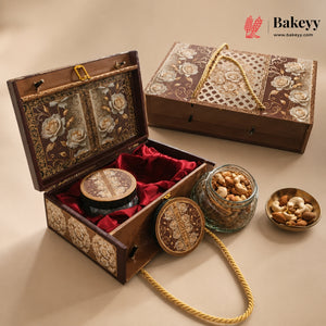An ornate wooden gift box set with floral inlay, red velvet lining, and gold rope handles, shown open with a round tin and jars of nuts beside it on a beige studio background. The Bakeyy logo appears in the top right.