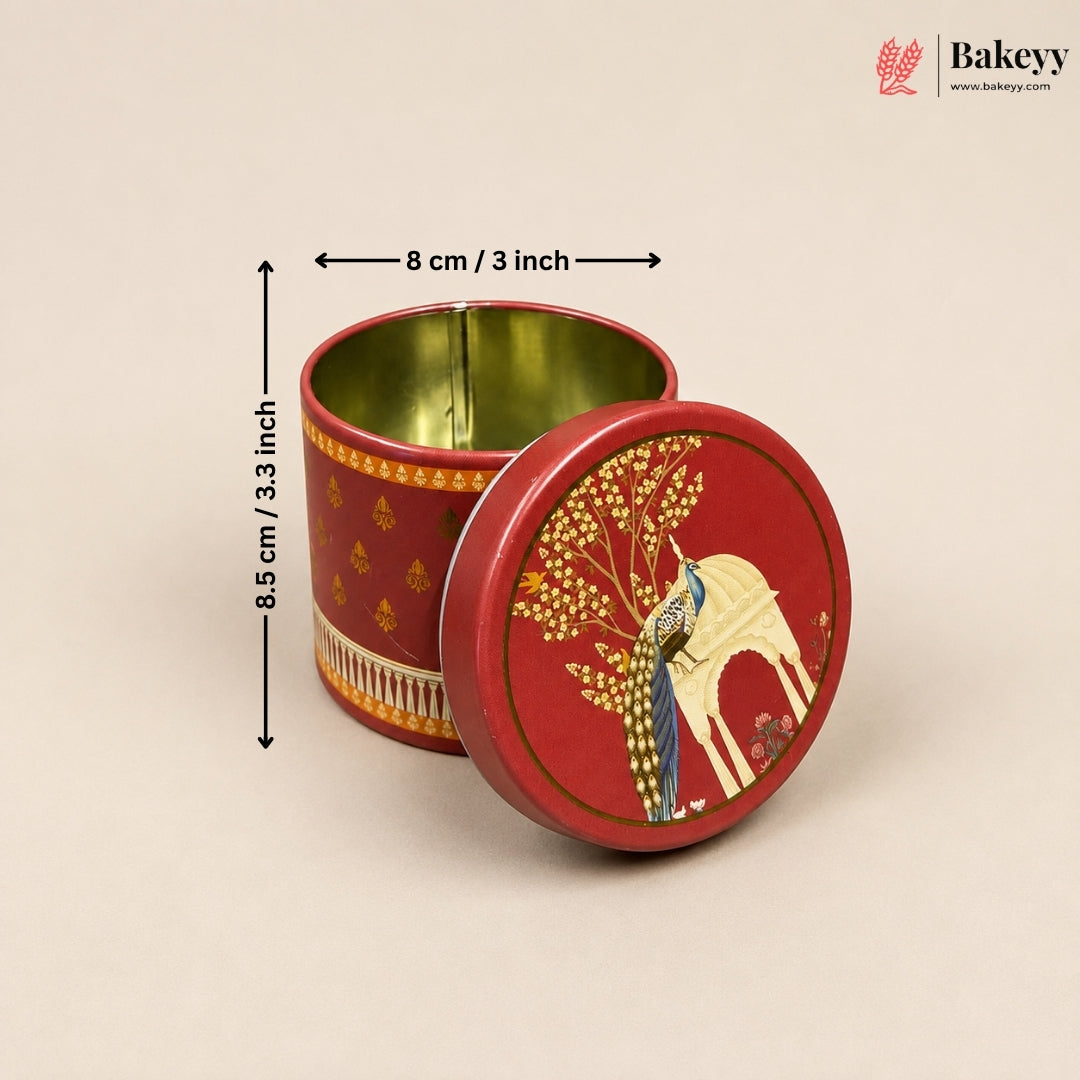 A round red metal tin with a matching lid, shown open on a light beige background. The lid features a gold and cream peacock and tree illustration, and the tin body has gold decorative patterns.