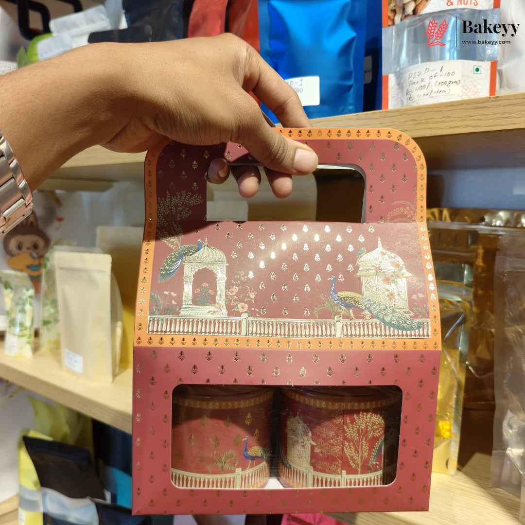 A hand holds a decorative red gift box with a handle, containing two matching red cylindrical tins with printed illustrations. The setting appears to be a retail shelf with other products in the background.