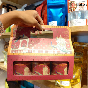 A hand holds a pink decorative gift box containing three red cylindrical tins with peacock and architectural illustrations, photographed on a store shelf.