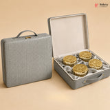 A gray textured hard case shown closed on the left and open on the right, containing four round gold-toned decorative items on a beige satin lining. The scene is photographed on a warm beige studio background with a Bakeyy watermark in the top right.