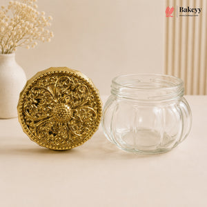 An empty clear glass jar with a decorative round gold lid placed beside it on a beige surface, with a vase of dried flowers and a vertical ribbed panel in the background.