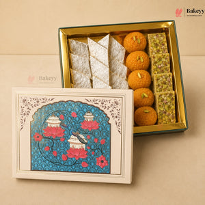 An open decorative gift box of Indian sweets and snacks on a beige background, with the illustrated lid placed in front. The box contains orange gulab halwa, rectangular pistachio-filled bars, and folded silver-colored sweets.