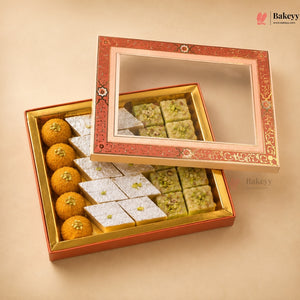 An open decorative gift box of Indian sweets on a beige background, with the lid leaning against the box. The box is red and gold with a clear window.