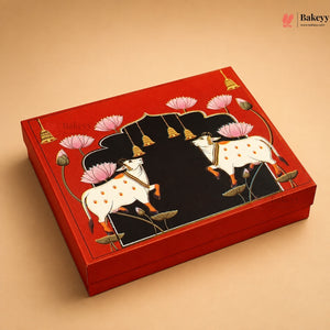 A red rectangular gift box with a hand-painted Madras-style illustration of two white cows with pink lotus flowers and a black central arch, shown on a warm beige background.