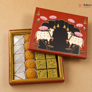 An open red and gold gift box of Indian sweets on a beige background, with the lid propped above showing a decorative illustration of two white cows under pink lotus flowers. The box contains assorted sweets, including orange marzipan, green pistachio-filled bars, and white sugar cubes.