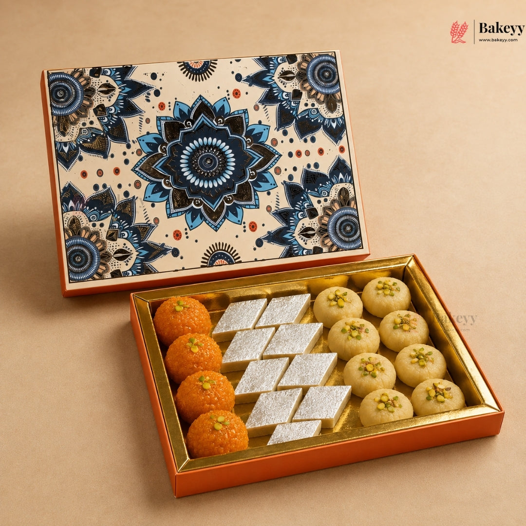 An open decorative gift box of Indian sweets on a beige background. The box has a cream lid with a blue mandala pattern and orange trim, and the tray inside is lined in gold.