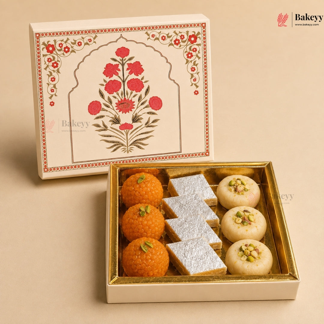 An open gold-trimmed gift box of Indian sweets on a beige background, with the matching illustrated lid leaning behind it. The box contains orange laddoo, white sugar-coated pieces, and cream-colored pistachio-topped sweets.