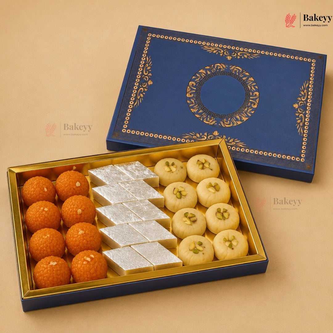 An open blue and gold gift box of Indian sweets on a beige background, with the matching lid placed behind it. The box contains orange marzipan laddons, white rectangular sweets, and round offal sweets with green pistachio centers.