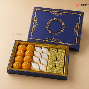 An open blue and gold gift box of traditional sweets on a beige background, with the matching lid placed behind it. The box contains orange round sweets, white square sweets, and green patterned sweets.