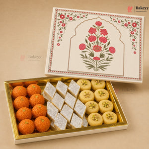An open gold-trimmed gift box of Indian sweets on a beige background, showing orange laddoo-style pieces, square white sugar-dusted cubes, and round yellow糕点. The lid is placed above and features a cream-colored floral arch design with red flowers and green leaves.
