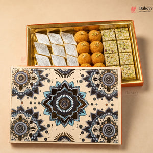 A boxed assortment of Indian sweets shown on a beige background, with the decorative lid in front and the open box behind. The open box contains silver-fried pastry pieces, round golden-brown laddons, and rectangular green-spiced bars.