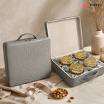 A gray textured hard case shown closed beside an open case displaying four round gold-lidded pastries or cookies on a cream satin insert. The scene is styled on a marble surface with beige fabric, a small dish of nuts, and dried flowers in a vase.