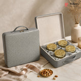 A gray textured hard case shown closed beside an open case displaying four round gold-lidded pastries or cookies on a cream satin insert. The scene is styled on a marble surface with beige fabric, a small dish of nuts, and dried flowers in a vase.