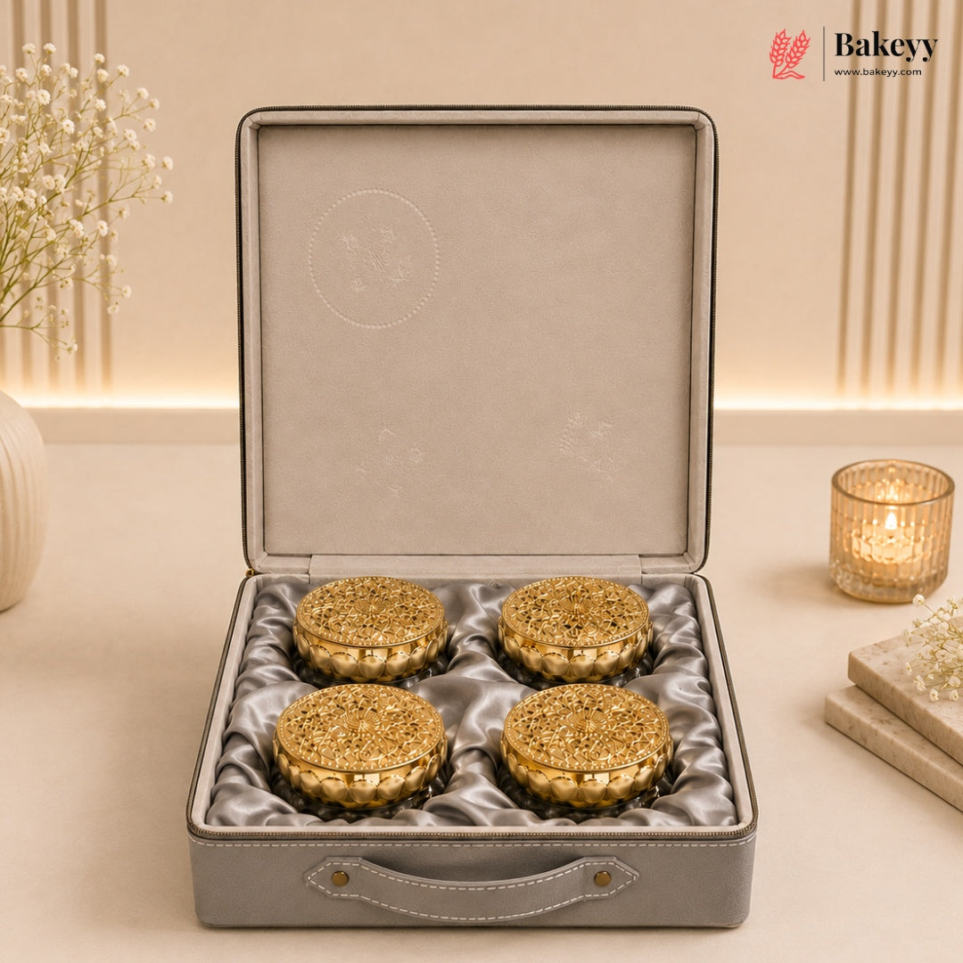 Four round golden-brown pastries arranged in a gray presentation box with a satin lining, photographed on a beige tabletop with decorative flowers and a lit candle in the background. The Bakeyy logo appears in the top right corner.