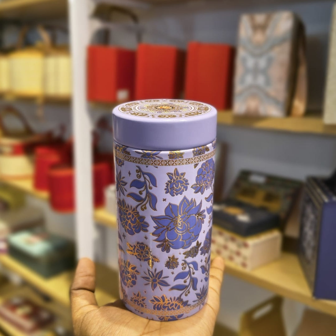 A lavender cylindrical tin with a blue and gold floral pattern, held in a hand in front of a blurred store background.
