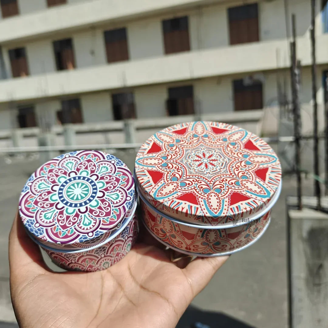 Hand holding two round tin boxes with colorful mandala prints, ideal for gift packaging from Bakeyy.com