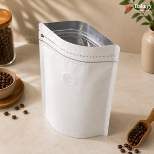 A white stand-up resealable coffee bag with a silver zipper, styled on a beige tabletop with coffee beans, a wooden scoop, and a small plant in the background.