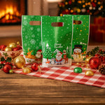 Three green Christmas gift bags with festive designs on a wooden table with holiday decorations.