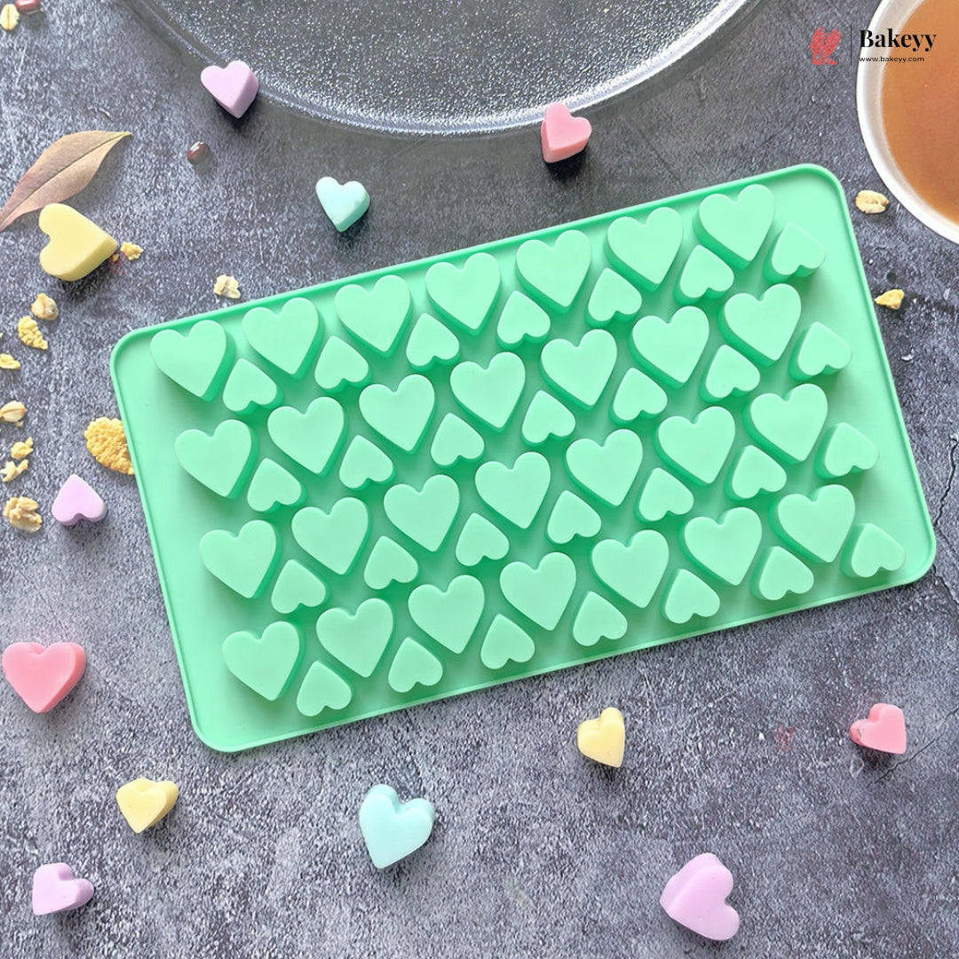 A mint green silicone mold tray with multiple heart-shaped cavities, shown on a gray textured surface with scattered pastel heart candies. A glass plate and a cup are partially visible at the top, and a Bakeey logo watermark appears in the upper right corner.