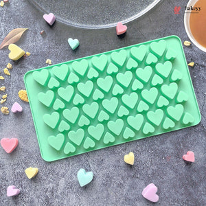 A mint green silicone mold tray with multiple heart-shaped cavities, shown on a gray textured surface with scattered pastel heart candies. A glass plate and a cup are partially visible at the top, and a Bakeey logo watermark appears in the upper right corner.