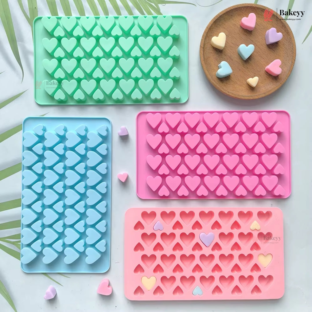 Four heart-shaped silicone baking molds in mint green, light blue, and pink, arranged on a white surface with a wooden tray holding small pastel heart-shaped candies. The Bakeyy brand logo is visible on the products.