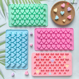 Four heart-shaped silicone baking molds in mint green, light blue, and pink, arranged on a white surface with a wooden tray holding small pastel heart-shaped candies. The Bakeyy brand logo is visible on the products.