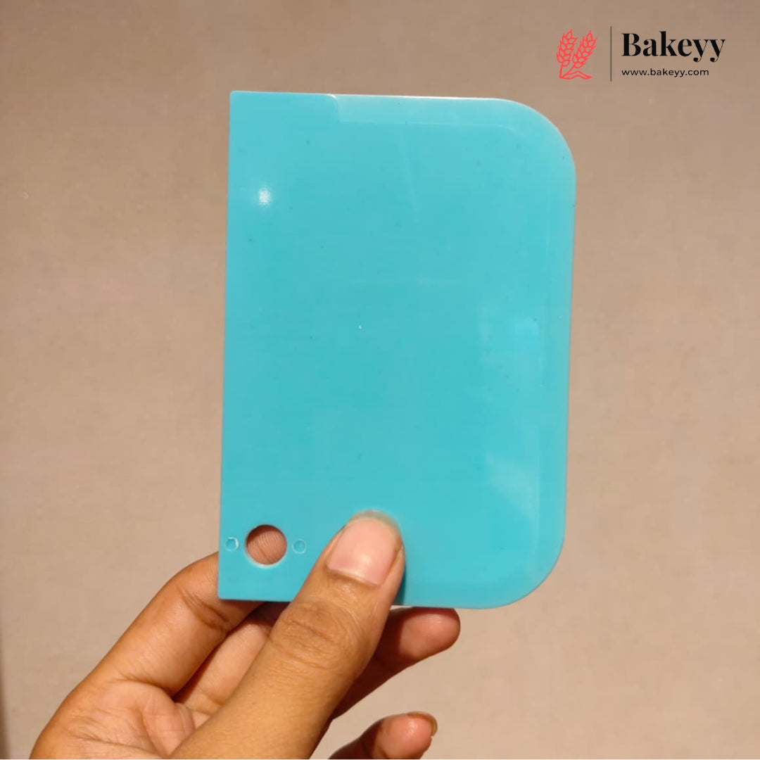 A hand holding a turquoise blue plastic scraper or spatula against a plain beige background, with a Bakeyy logo watermark in the top right corner.