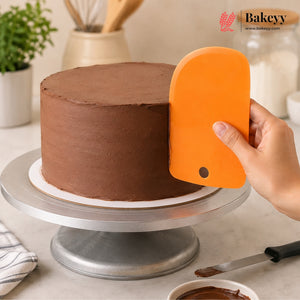 An orange cake scraper being used to smooth the side of a chocolate frosted cake on a silver rotating cake stand, with a hand holding the scraper. The Bakeyy logo is visible in the top right corner.
