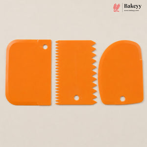 Three bright orange silicone pastry tools with different blade and scraper designs, arranged side-by-side on a light beige background. The Bakeyy logo is visible in the top right corner.