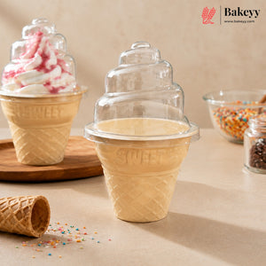 A clear plastic ice cream cone lid with a tiered swirl design sits on top of a waffle cone cup in the center. A second cone with a pink-and-white swirled dessert is behind it on a wooden board, with bowls of sprinkles and a spilled waffle cone in the background.