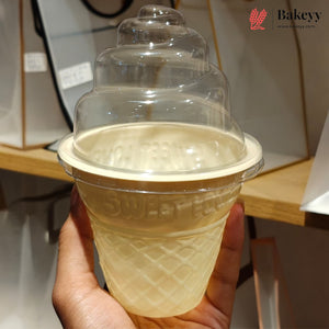 A hand holding a clear plastic ice cream cone-shaped lid with a beige waffle-textured base, set against a blurred indoor background with shelves and boxes. A Bakeyy logo is visible in the top right corner.