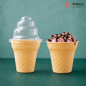 Two yellow waffle-cone-shaped dessert cups on a teal surface against a teal background. The left cup has a clear plastic tiered lid, and the right cup is filled with pink ice cream or gelato topped with chocolate drizzle and sprinkles.