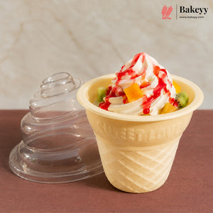 A beige waffle-cone-shaped dessert cup filled with whipped cream, fruit pieces, and red syrup, shown next to a clear plastic lid on a brown surface against a light stone background. The Bakeey logo appears in the top right.