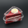 Slice of red velvet cake with white frosting in a clear plastic container on a black background