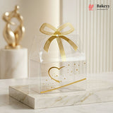 Clear gift box with gold bow and heart design on a marble surface, featuring the Bakey brand.