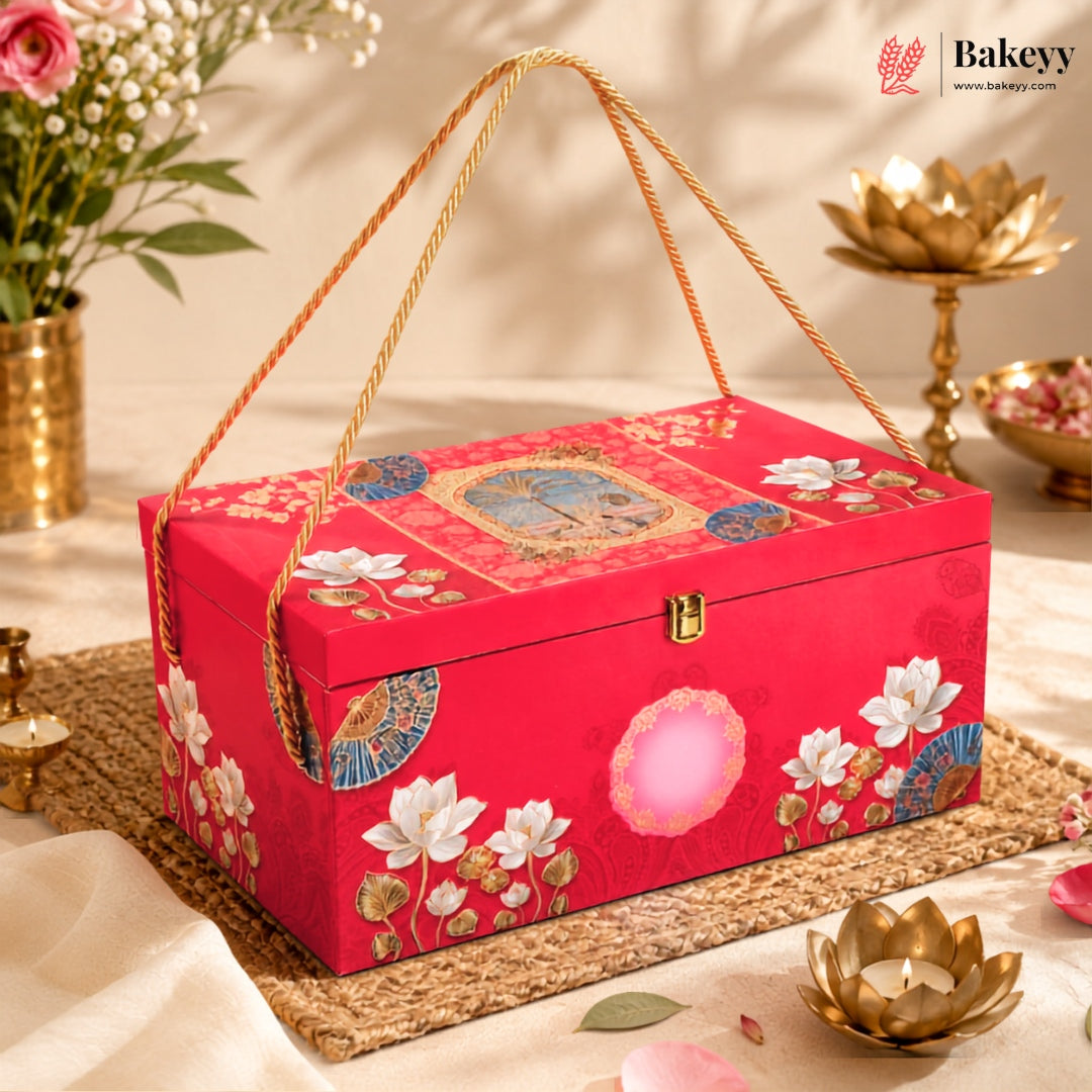 A red decorative gift box with floral and circular printed motifs, tied with a rope handle, displayed on a woven mat with gold lotus candle holders and flowers in the background.